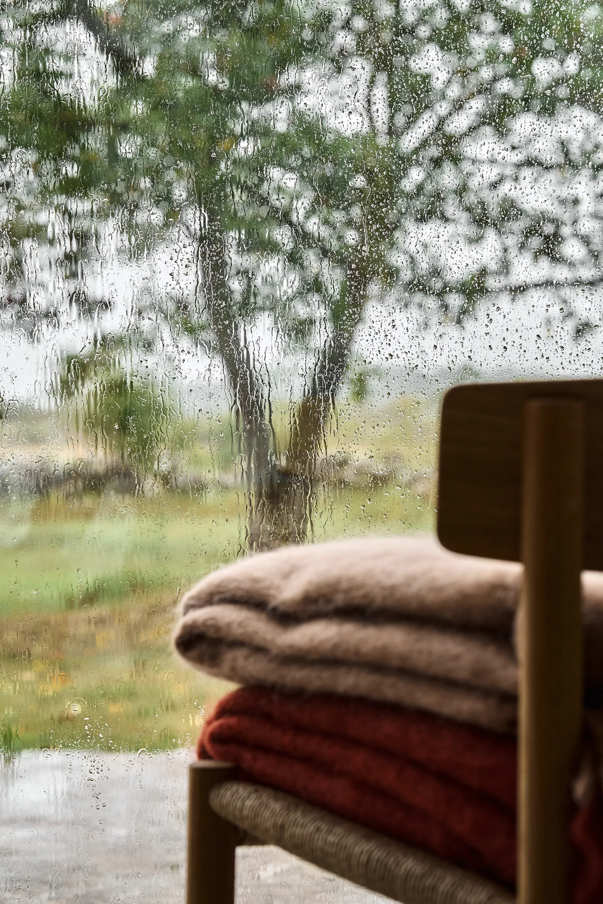 Mantas apiladas en una silla de madera junto a una ventana con gotas de lluvia y árboles borrosos.