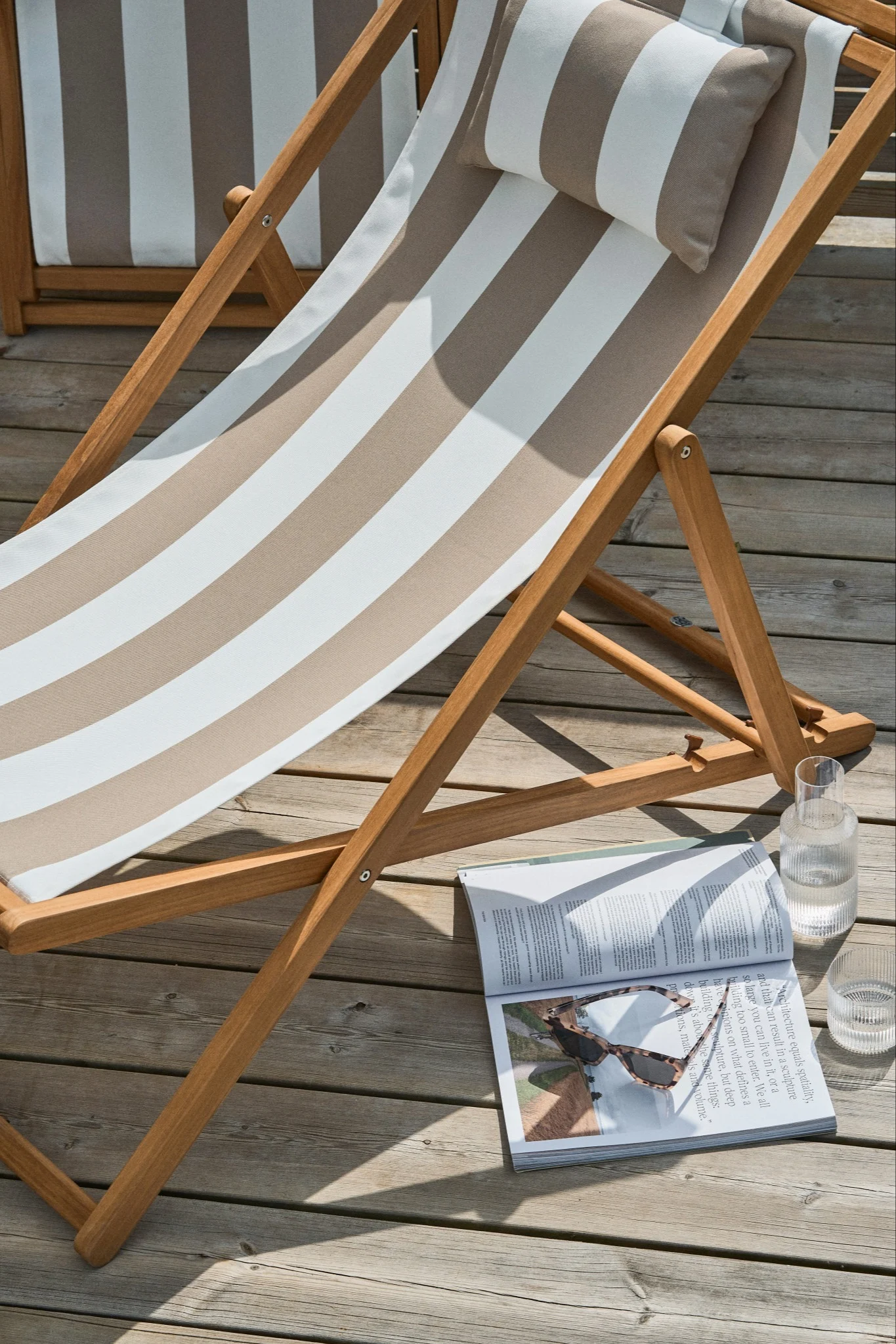 Una tumbona de madera con cojín y almohada a rayas, una revista con gafas de sol y un vaso de agua en una terraza de madera.