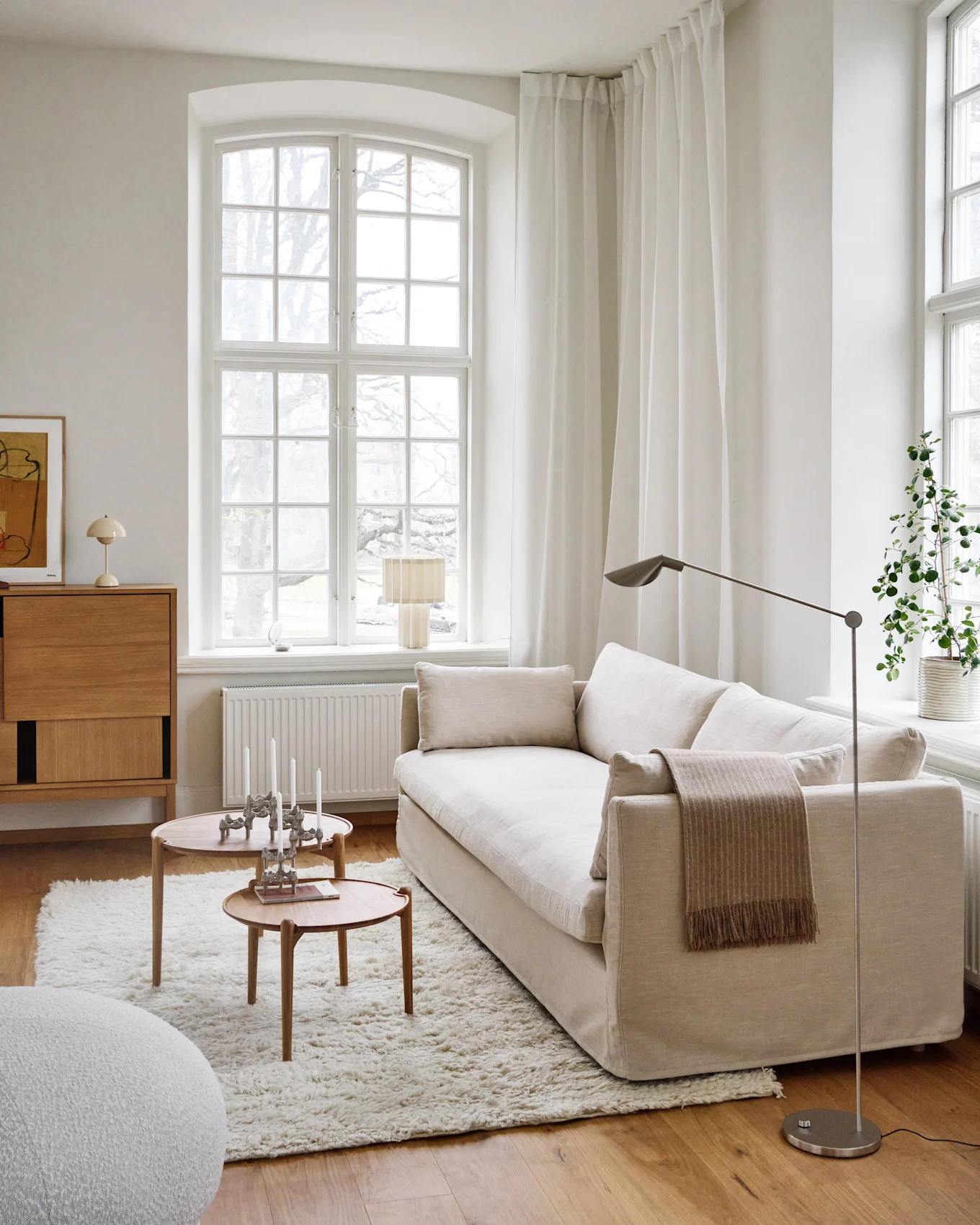 Una sala de estar luminosa y minimalista con un sofá beige claro, dos mesas de centro redondas de madera con velas, una alfombra blanca peluda y una ventana grande.
