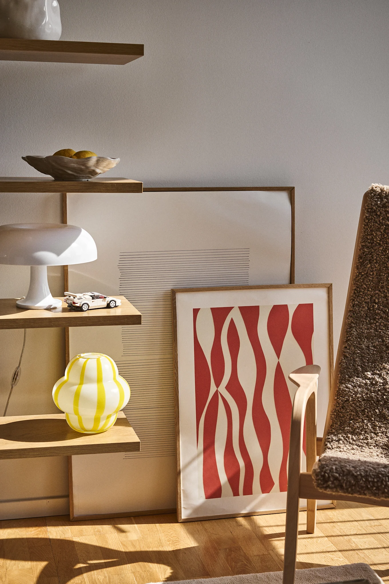 El jarrón Curlie de Byon tiene un diseño redondo y bulboso en vidrio a rayas amarillas y blancas. Aquí colocado en una estantería abierta junto con el cuenco Shell, las lámparas y los cuadros.