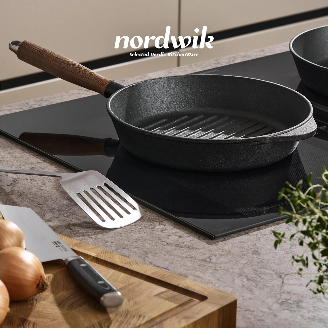 Una sartén grill de hierro fundido negra sobre una placa de inducción, con una espátula, cebollas, cuchillo y tomillo en una tabla de madera. Logo de Nordwik visible.