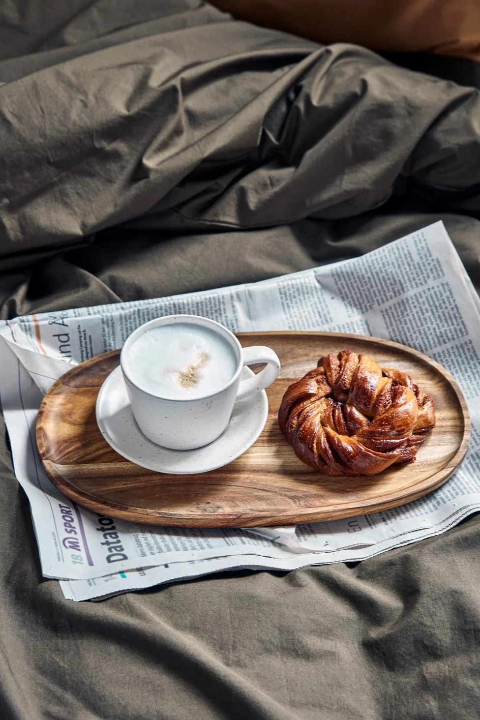 Un capuchino en la taza blanca y gris Pion de House Doctor en una pequeña bandeja de madera, sobre un periódico, en una cama, con un dulce sueco de canela al lado.