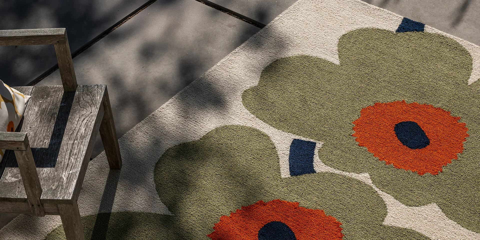 Alfombra exterior floral verde oliva y naranja junto a una silla de madera envejecida en un patio de hormigón.