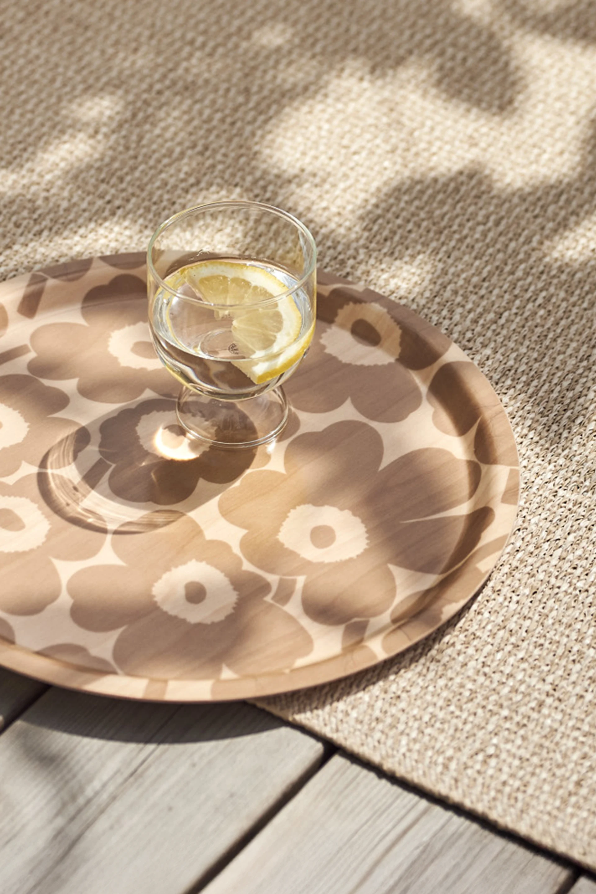 Un vaso de agua con rodaja de limón en una bandeja redonda estampada, sobre una alfombra tejida beige y tarima de madera con luz solar moteada.