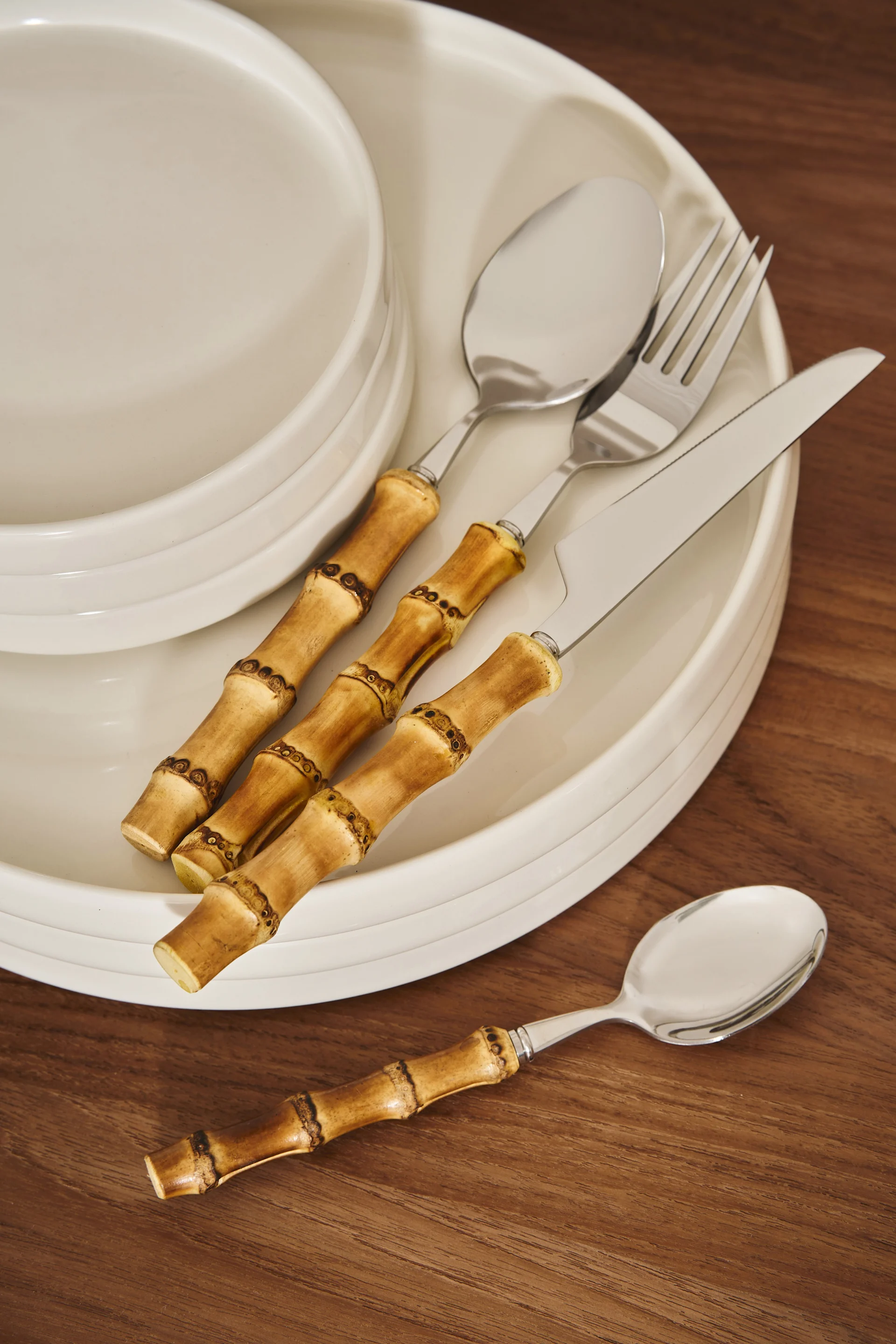 Juego de cubiertos con mango de bambú (cuchillo, tenedor, cucharas) sobre platos blancos y una mesa de madera.