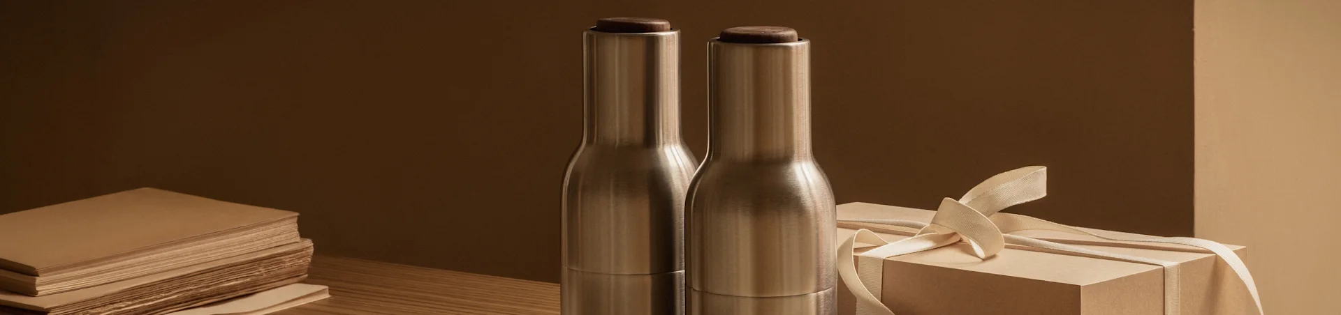 Dos botellas de metal cepillado con tapas de madera oscura junto a una caja de regalo marrón claro con un lazo sobre una mesa de madera.