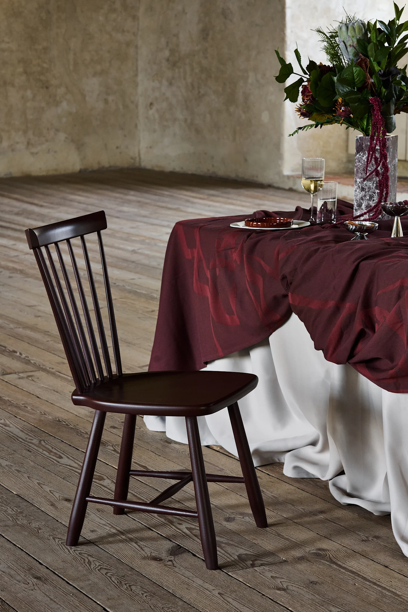 Silla de madera marrón oscuro con respaldo de listones, junto a una mesa cubierta de burdeos con un arreglo floral, sobre un suelo de madera.