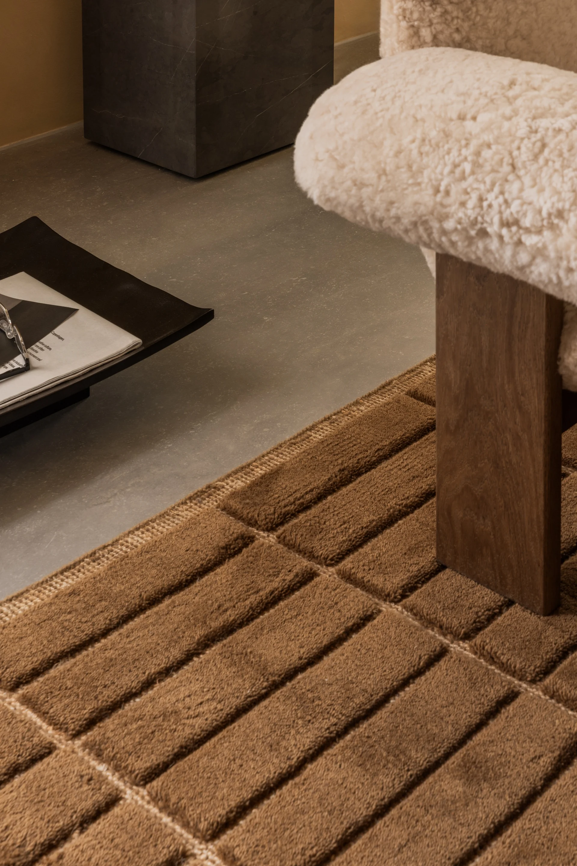 Alfombra marrón texturizada con patrones de bloques sobre un suelo de hormigón junto a una silla crema mullida y una mesa auxiliar oscura.