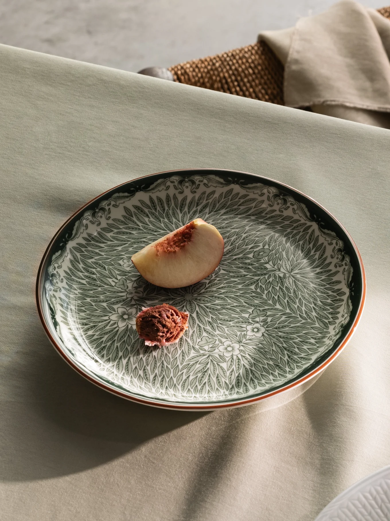 Una mesa puesta con un plato verde estampado con una rebanada de melocotón, y vista parcial de un plato blanco con crema y rebanadas de melocotón.