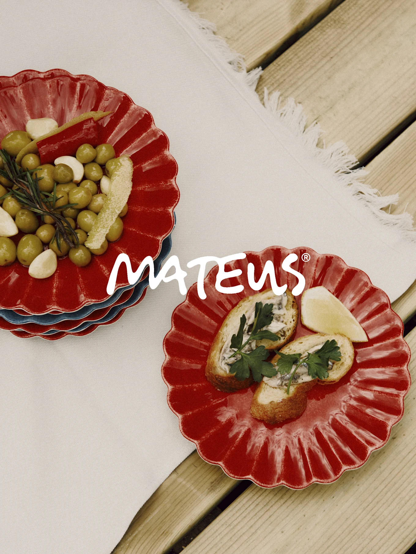 Platos rojos festoneados con aceitunas, ajo, romero y pan con anchoas en una mesa de madera. Marca MATEUS.
