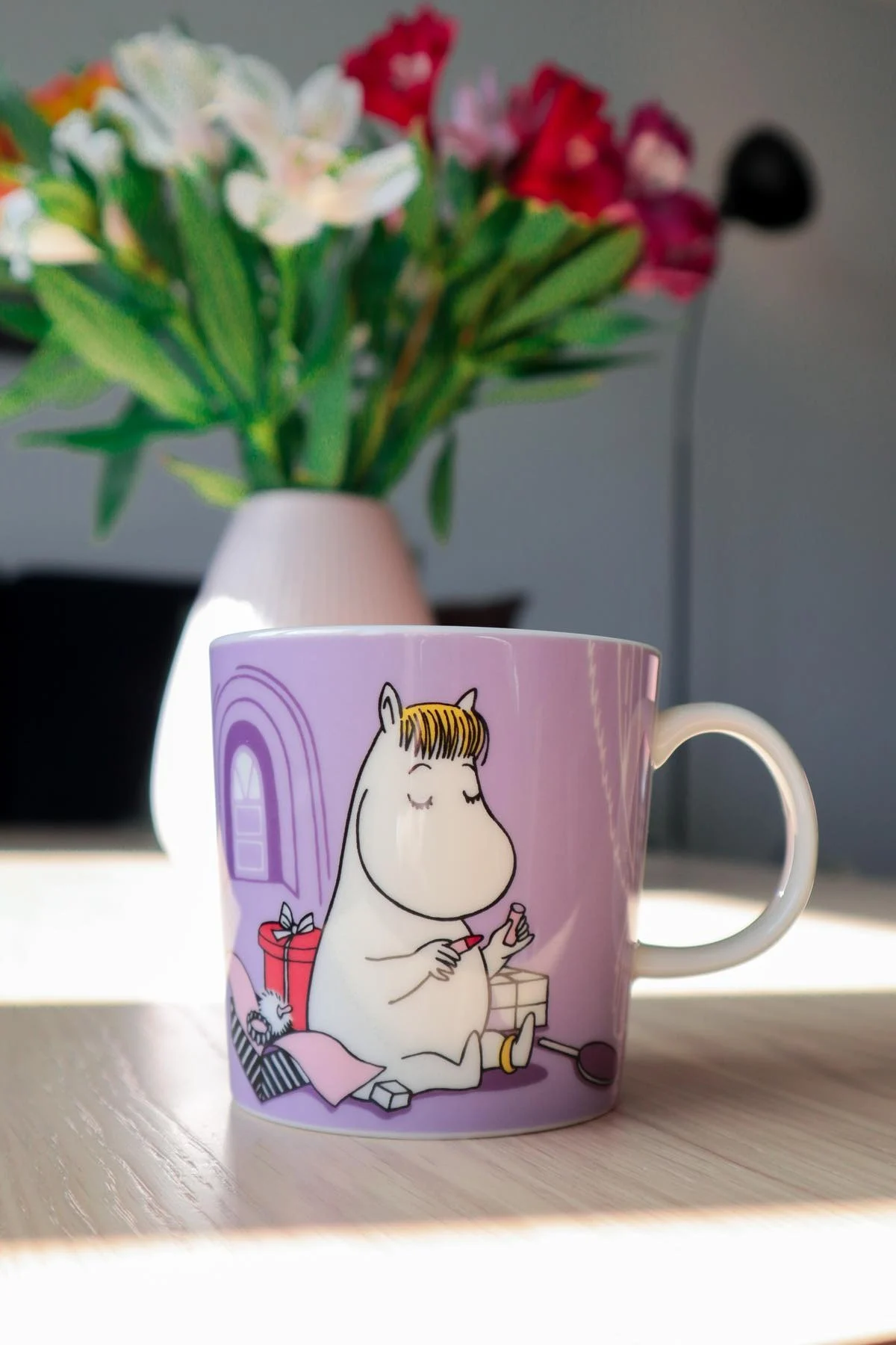 La taza morada de Moomin con la niña Snorky está sobre una mesa, al fondo hay un jarrón con un ramo de flores.