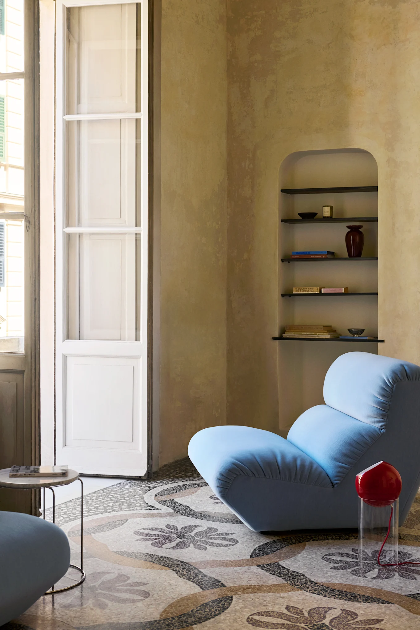 Un sillón azul claro sobre un suelo de mosaico estampado junto a una lámpara roja, con un estante arqueado y una ventana alta.