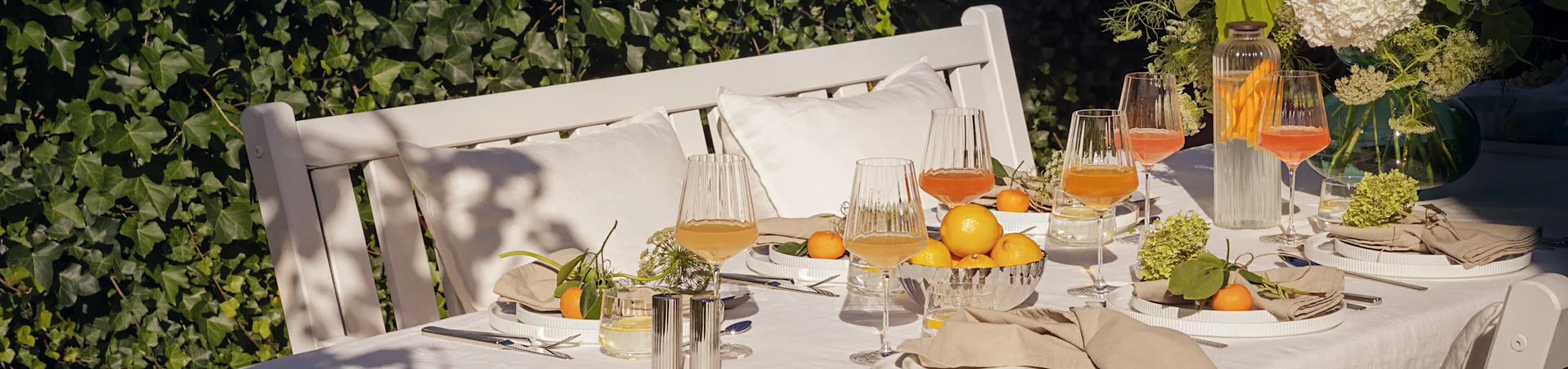 Mesa exterior para una comida de verano con platos blancos estriados, servilletas de lino, vasos estriados con bebidas, limones y mandarinas.