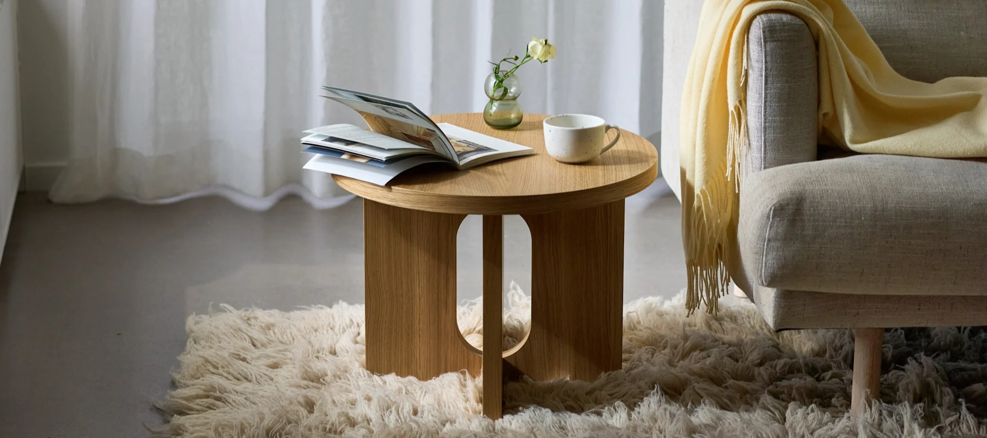 Mesa auxiliar redonda de madera con revista, jarrón y taza, junto a un sillón con manta amarilla sobre alfombra mullida.