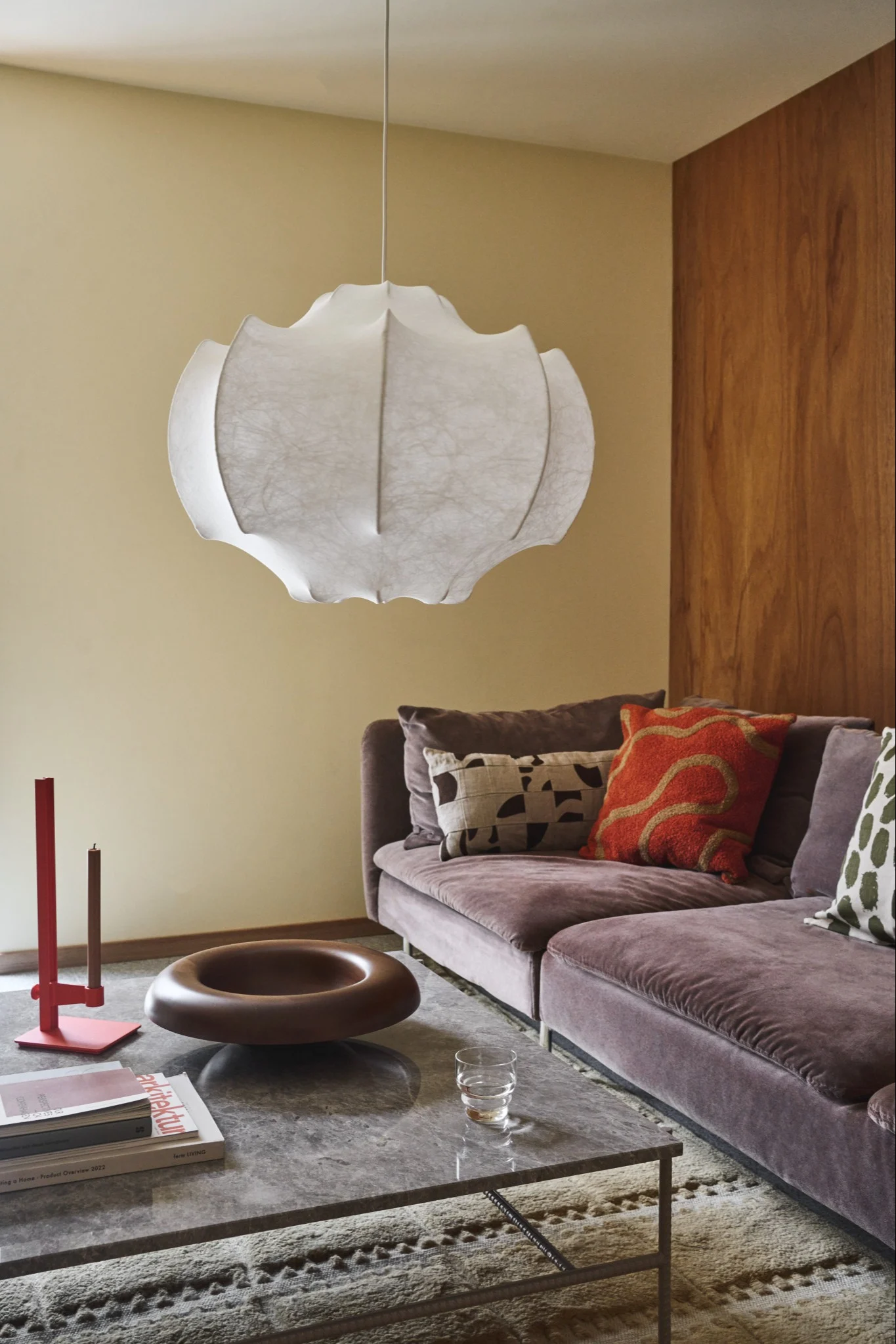 Un salón con una lámpara de papel blanca y ondulada, un sofá de terciopelo ciruela, una mesa de centro de mármol gris y elementos decorativos.