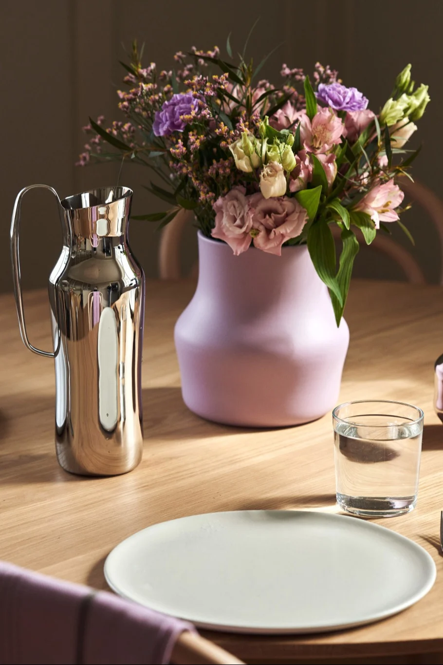 Aquí puedes ver el jarrón Dorotea morado con un ramo de flores rosas y moradas sobre una mesa junto a la jarra Dorotea plateada.