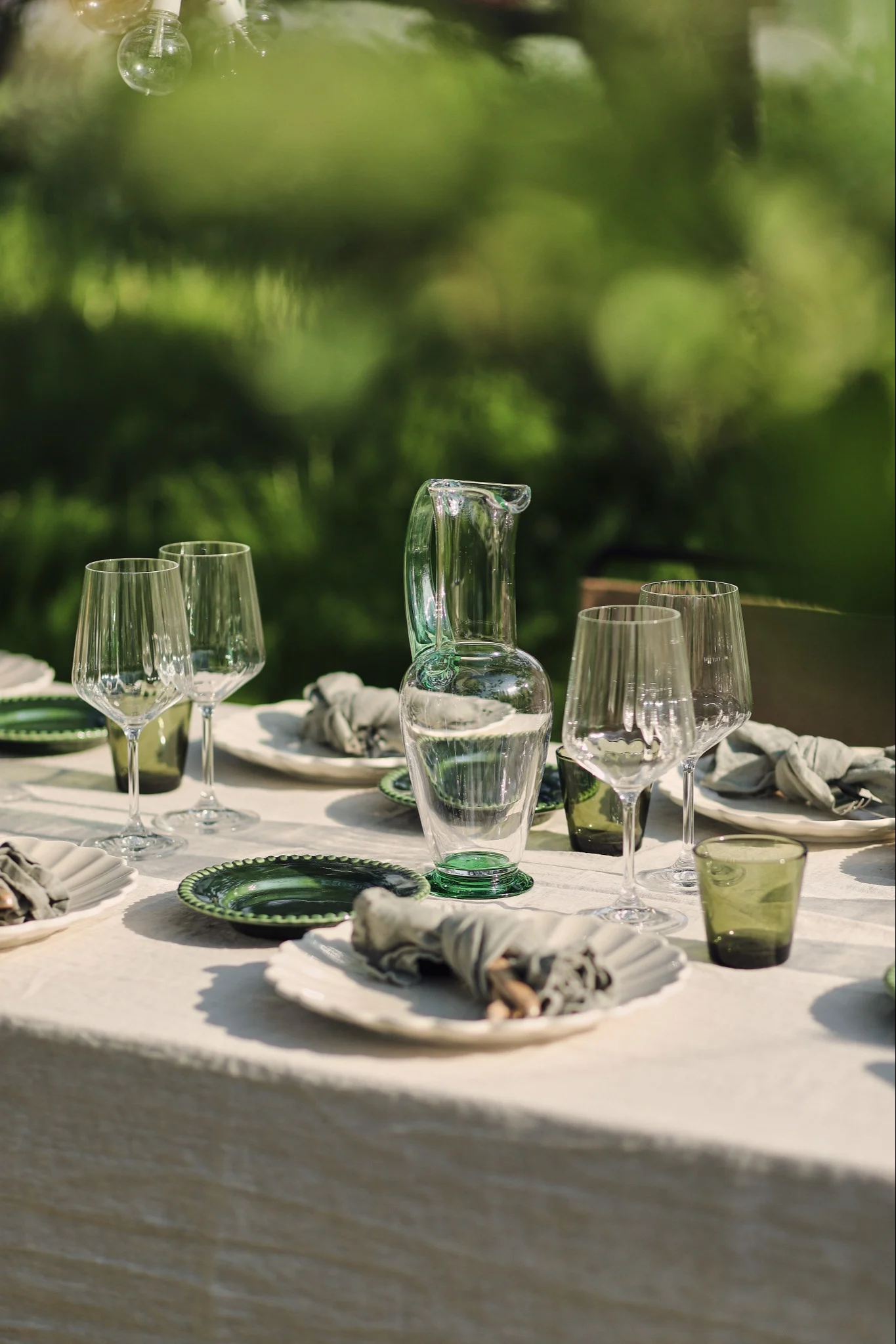 Elegante mesa de exterior con platos verdes y blancos, copas de vino transparentes y una jarra con base verde sobre un mantel blanco.