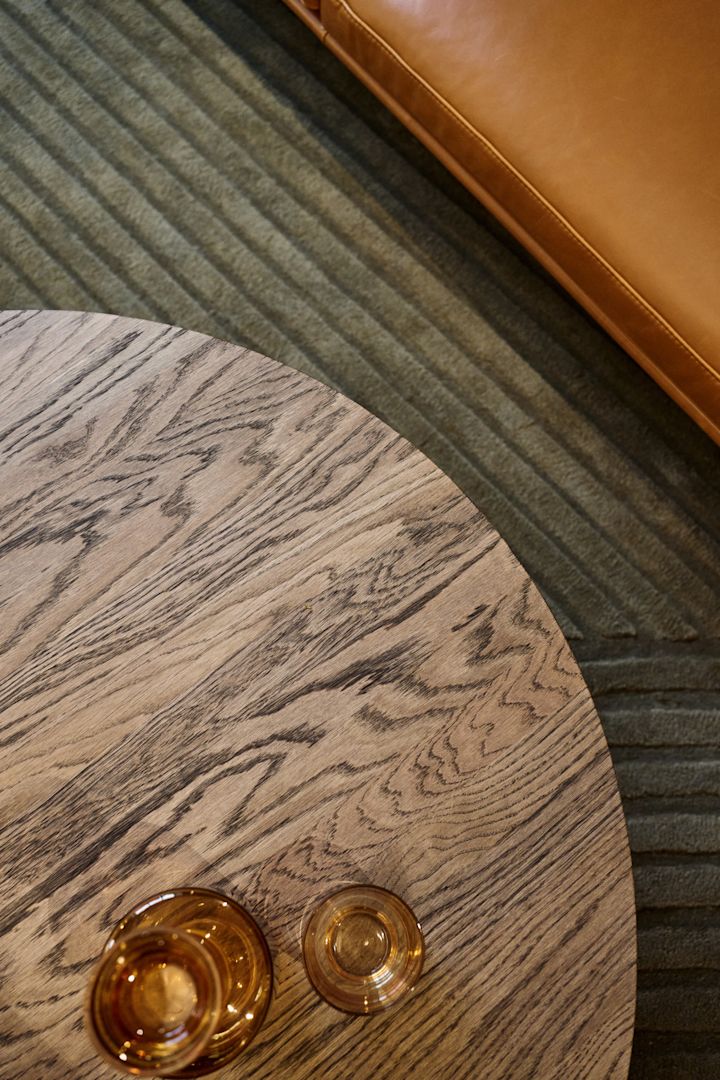 Una mesa de centro con tablero marrón oscuro se alza frente a un sofá, con varios vasos de color ámbar de la colección Raise de Muuto encima.
