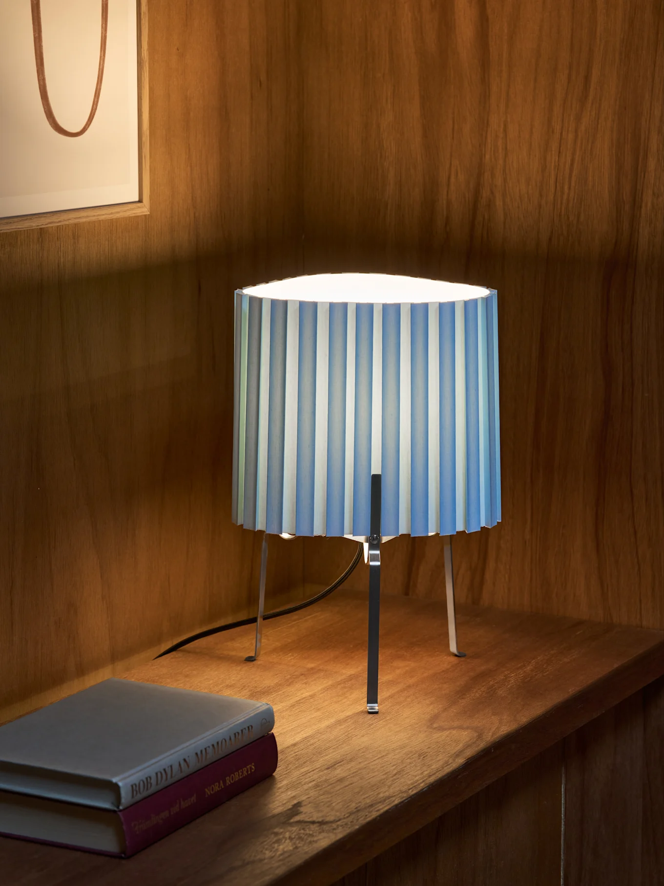 Lámpara de mesa plisada a rayas azules y blancas con tres patas metálicas sobre un estante de madera junto a dos libros. Obra de arte enmarcada detrás.
