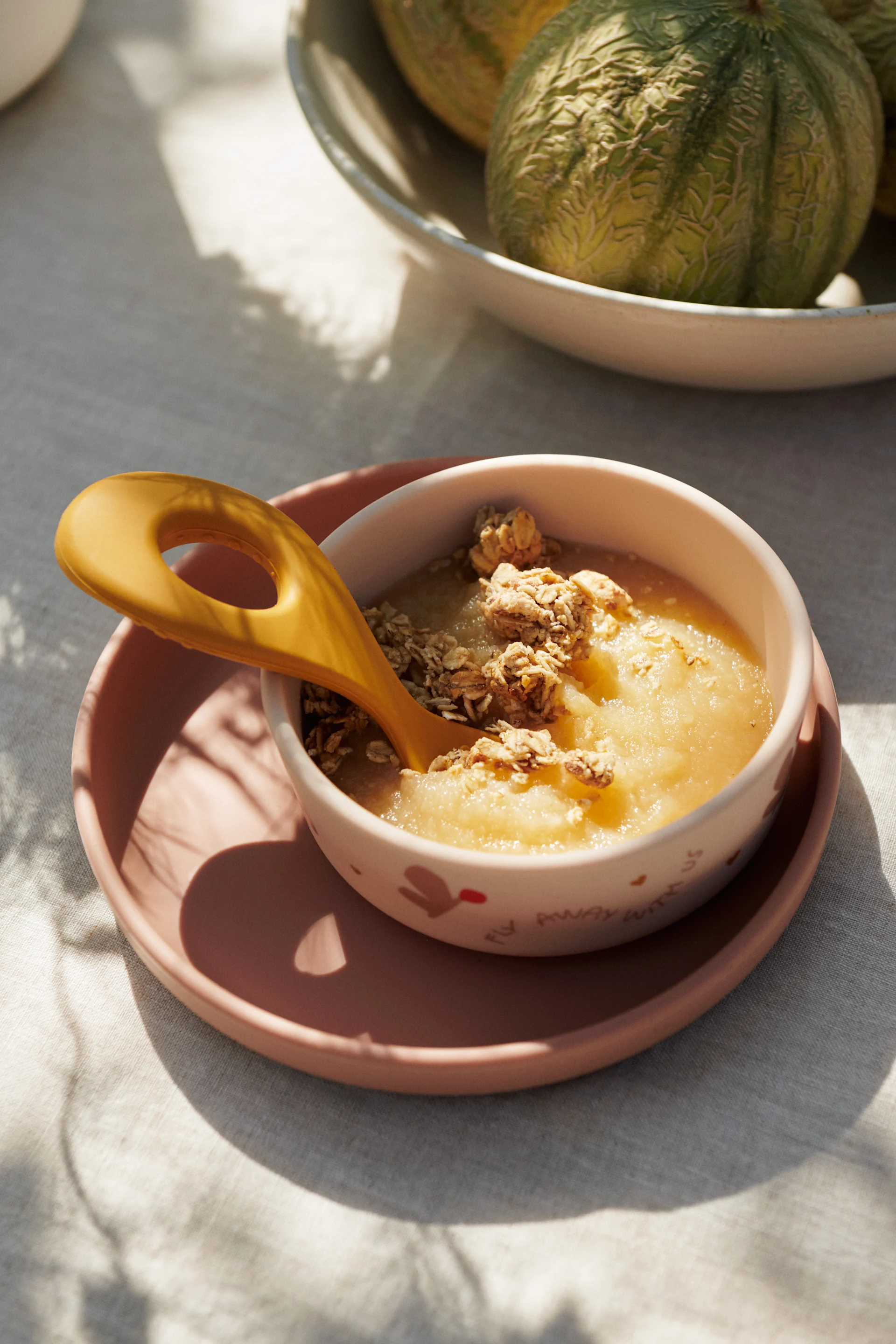 Puré de manzana con granola y una cuchara amarilla en un tazón sobre un plato rosa, con melones.