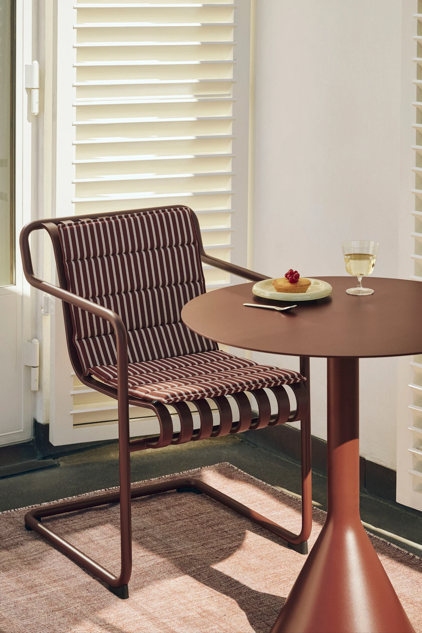 Una silla marrón rojiza con cojín a rayas junto a una mesa redonda a juego con un pastel y una copa de vino blanco.