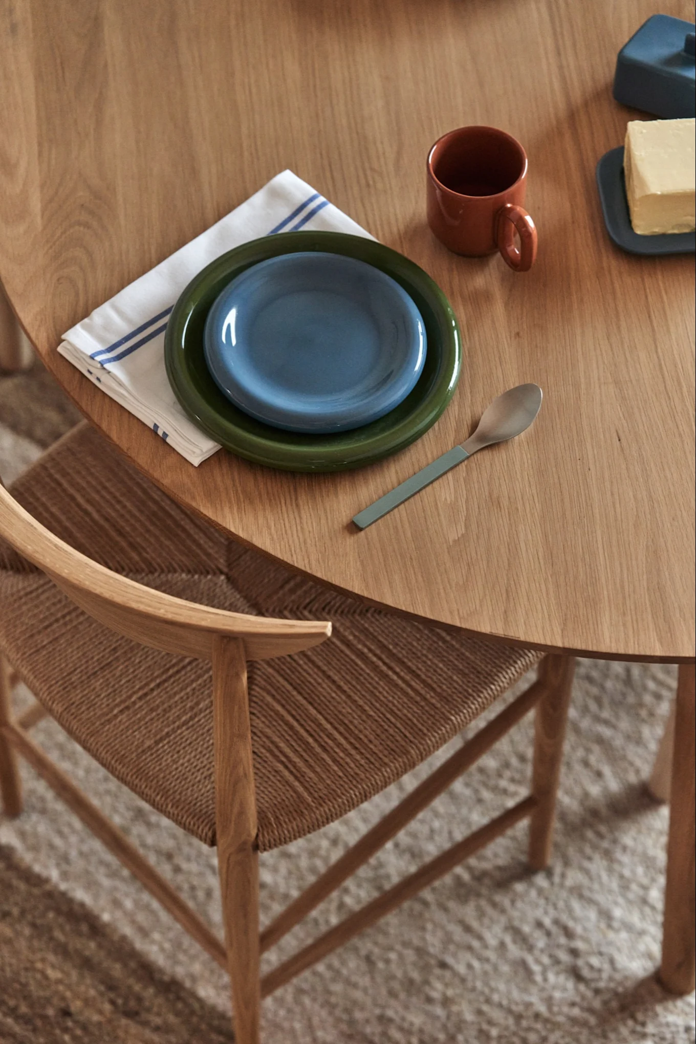 Una mesa de madera con un servicio de mesa, incluyendo platos verdes y azules, una servilleta rayada, una taza, una cuchara y mantequilla. Una silla tejida es visible.
