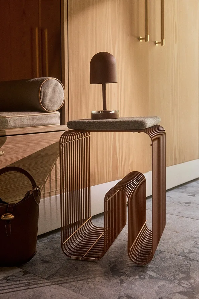 Mesa auxiliar moderna de varillas metálicas verticales marrones con una lámpara de cúpula marrón y un bolso, frente a un armario de madera.