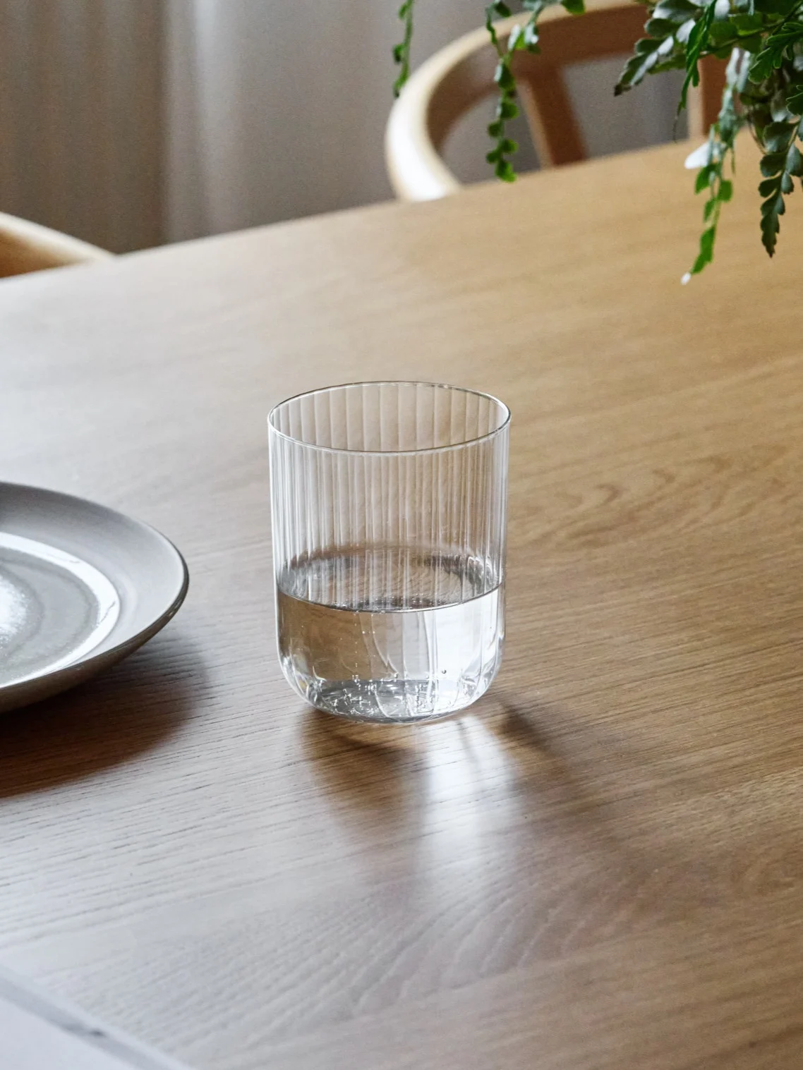 Un vaso de vidrio acanalado, medio lleno de agua, sobre una mesa de madera junto a un plato de cerámica.