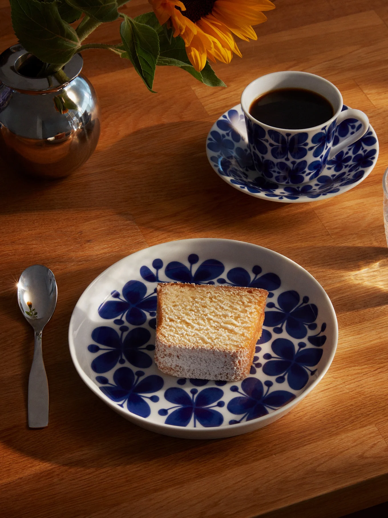 Una rebanada de pastel en un plato floral azul y blanco, con una taza de café, un vaso de agua, una cuchara y un girasol en un jarrón plateado sobre una mesa de madera soleada.