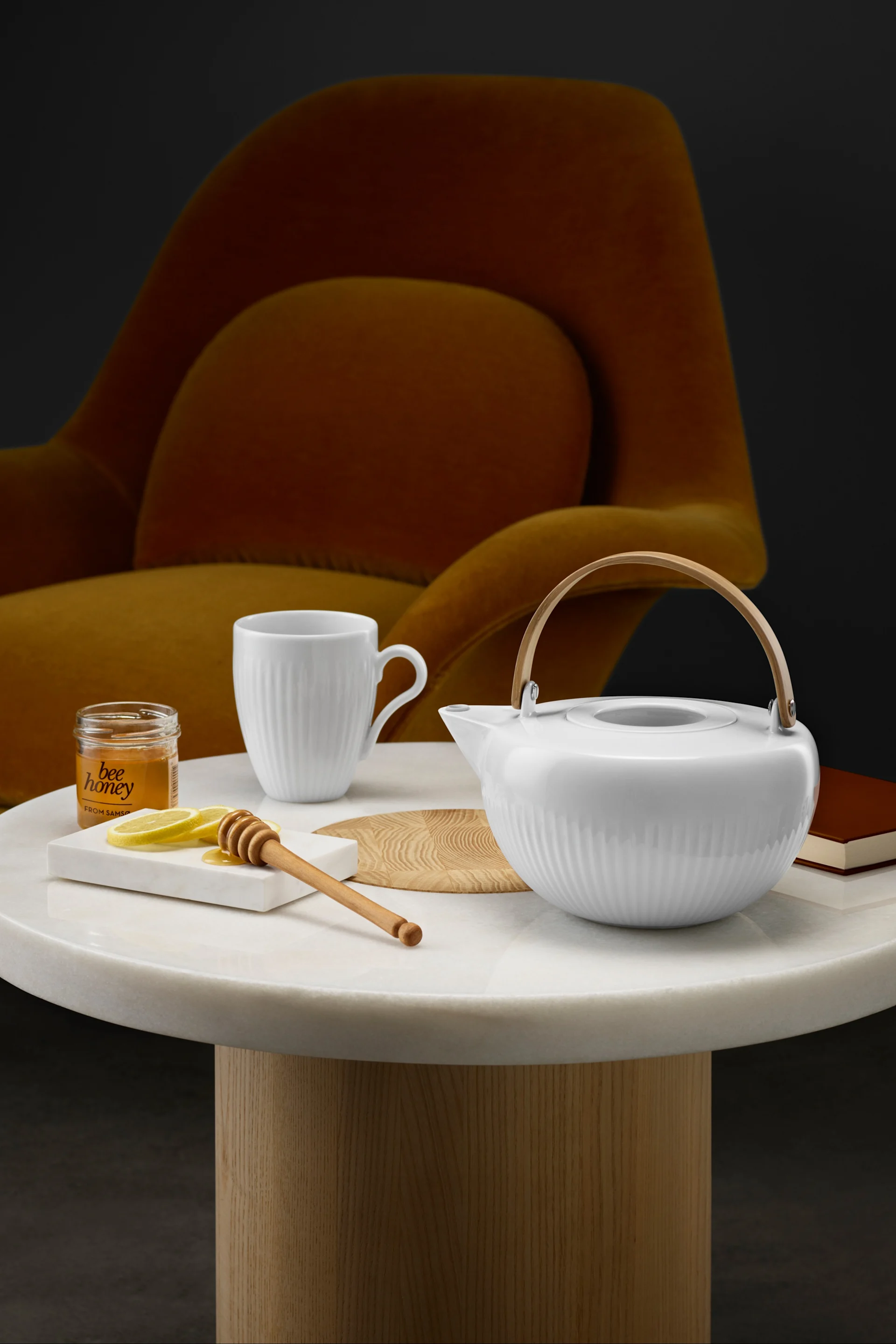 Tetera y taza blancas acanaladas, tarro de miel, rodajas de limón y cuchara para miel en una mesa de mármol con una silla naranja al fondo.