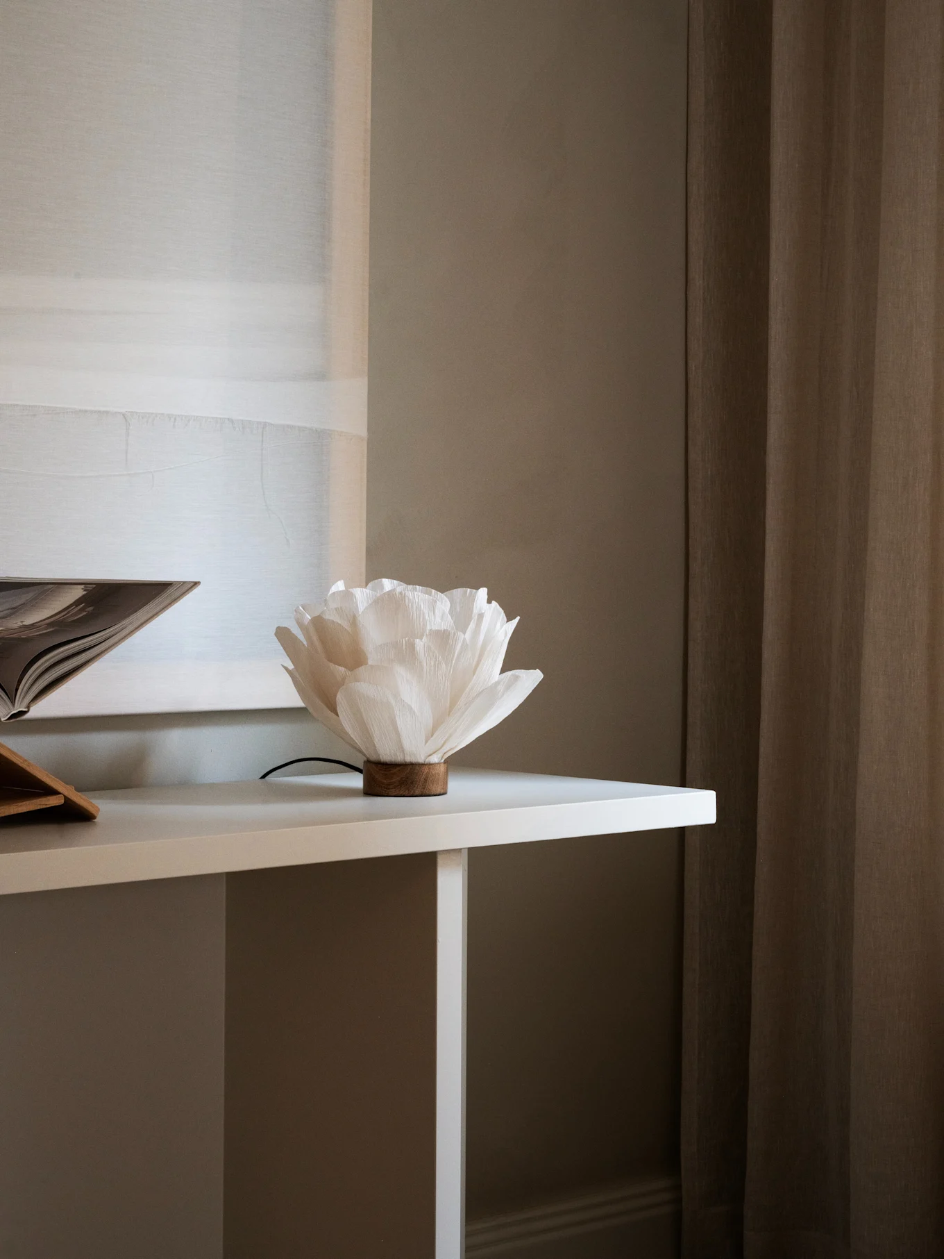 Lámpara de papel blanca con forma de flor y base de madera sobre una mesa blanca, junto a un libro abierto. Cortinas marrones al fondo.