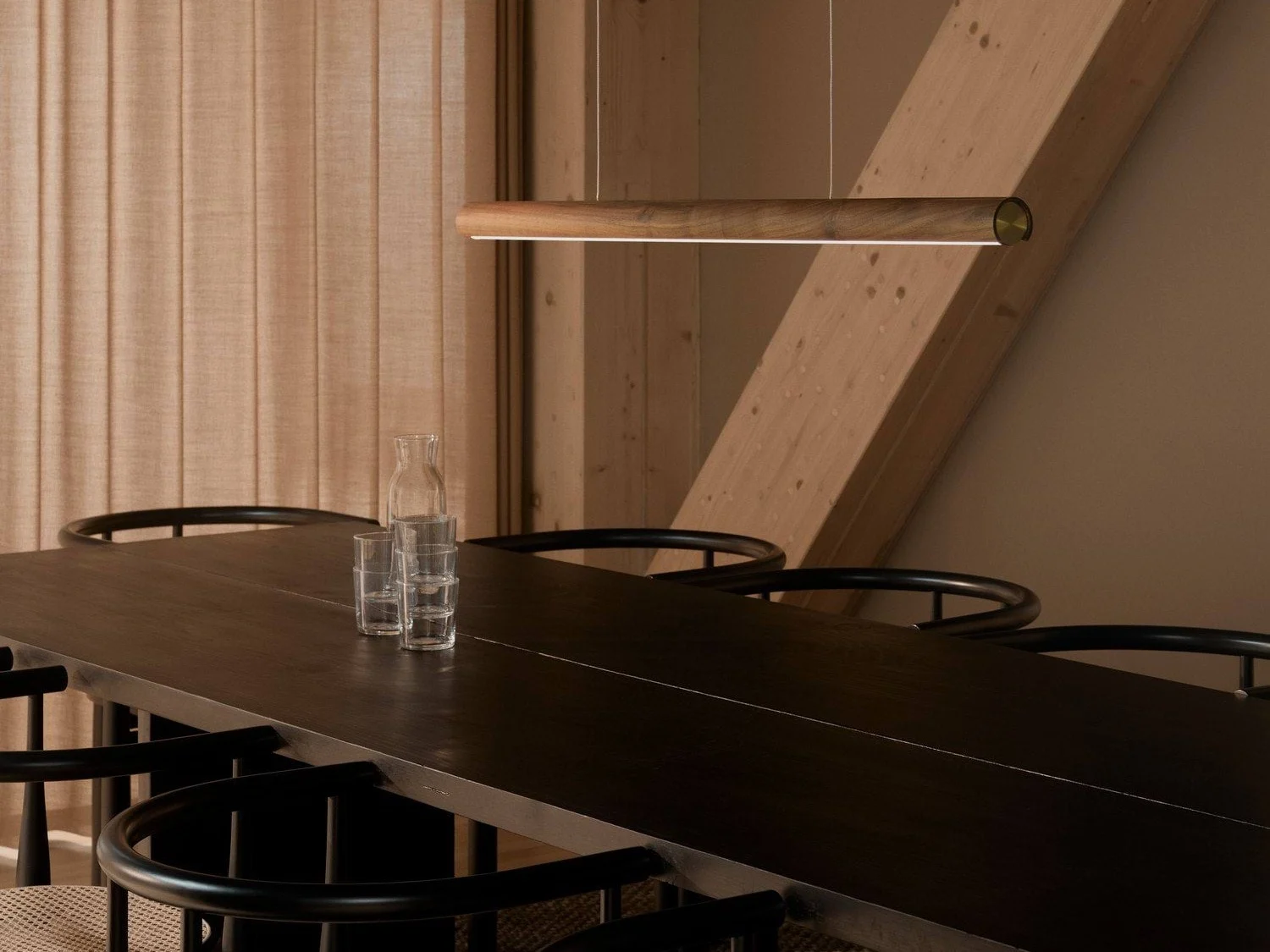 Un comedor moderno con una mesa larga de madera oscura, sillas negras y una elegante lámpara colgante de madera. Una jarra de vidrio y tres vasos están sobre la mesa.