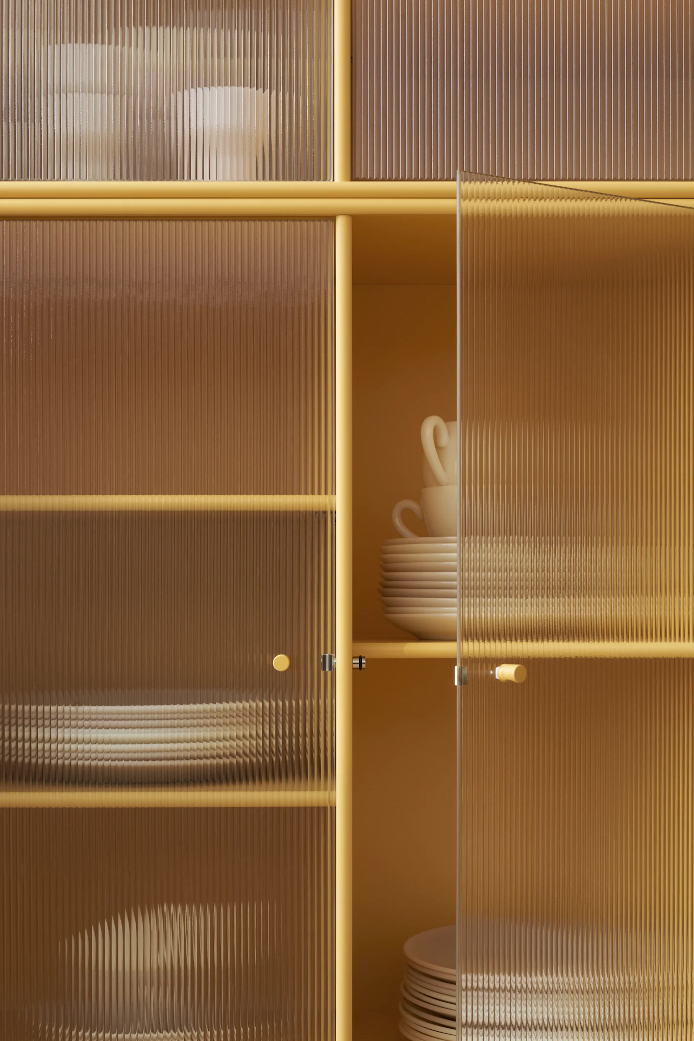 Armario amarillo mostaza con puertas de cristal estriado, revelando platos y tazas blancas apiladas en el interior. Una puerta está entreabierta.