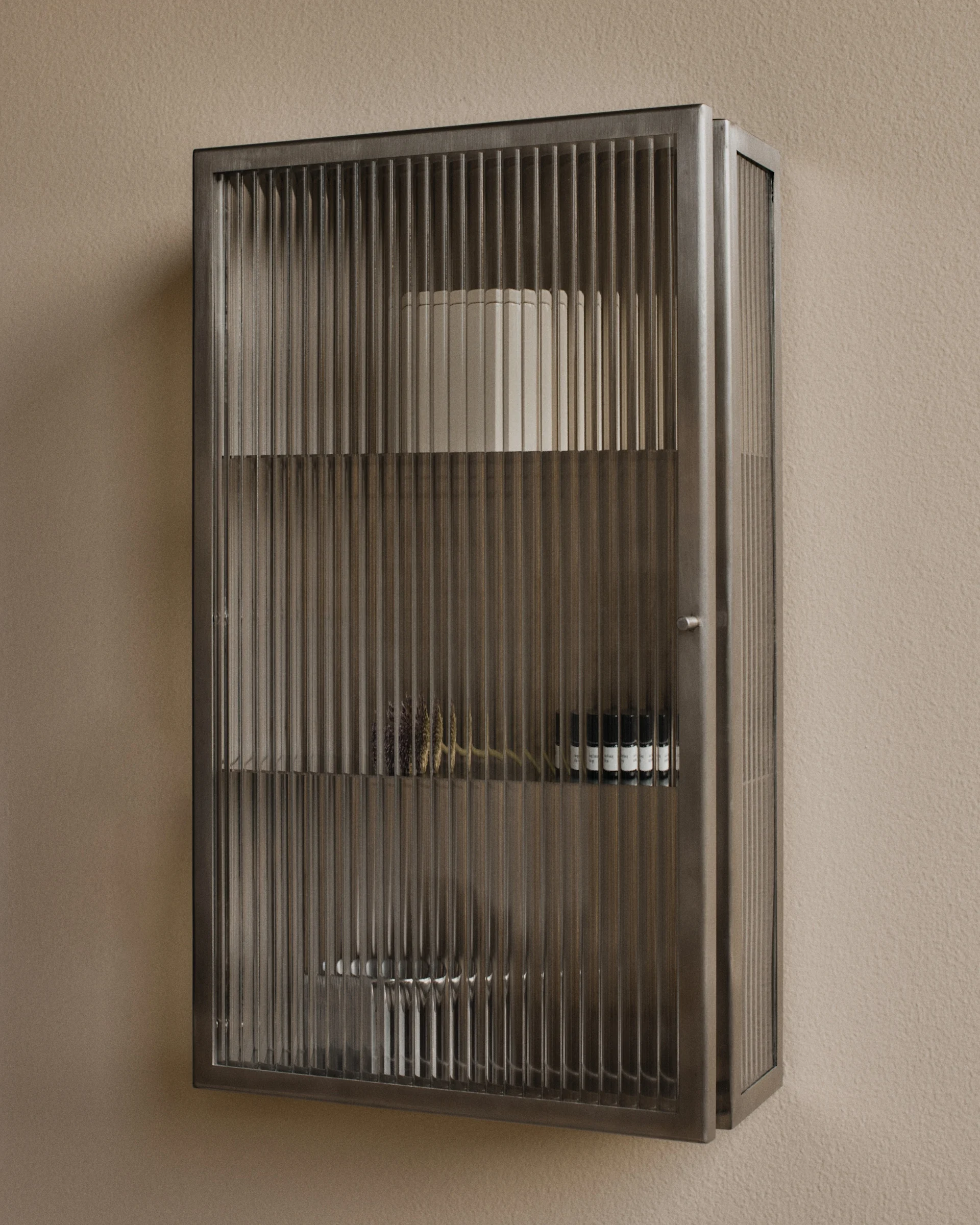 Armario metálico de pared con puertas de cristal estriado, que contiene objetos decorativos y pequeños frascos.