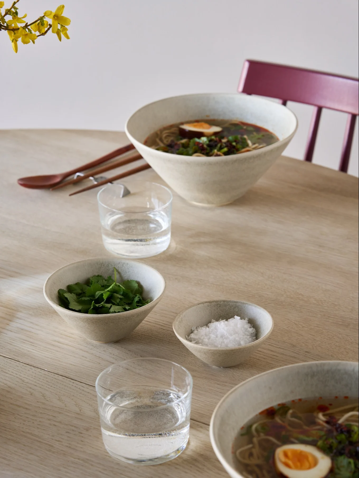 Dos tazones de ramen con huevos y hierbas, tazones de acompañamiento con cilantro y sal, agua, palillos y una cuchara en una mesa de madera clara.