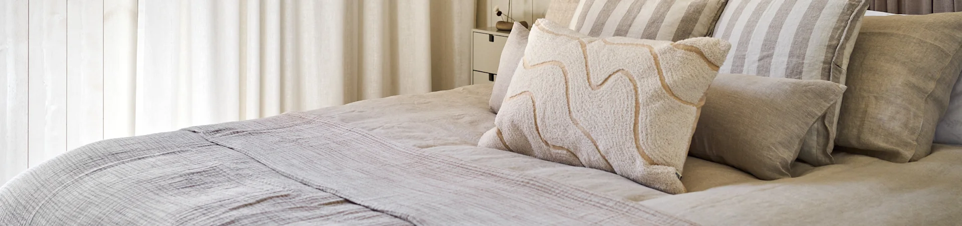 Una cama con ropa de cama de lino de tonos neutros, almohadas a rayas y una almohada blanca texturizada con líneas beige onduladas.