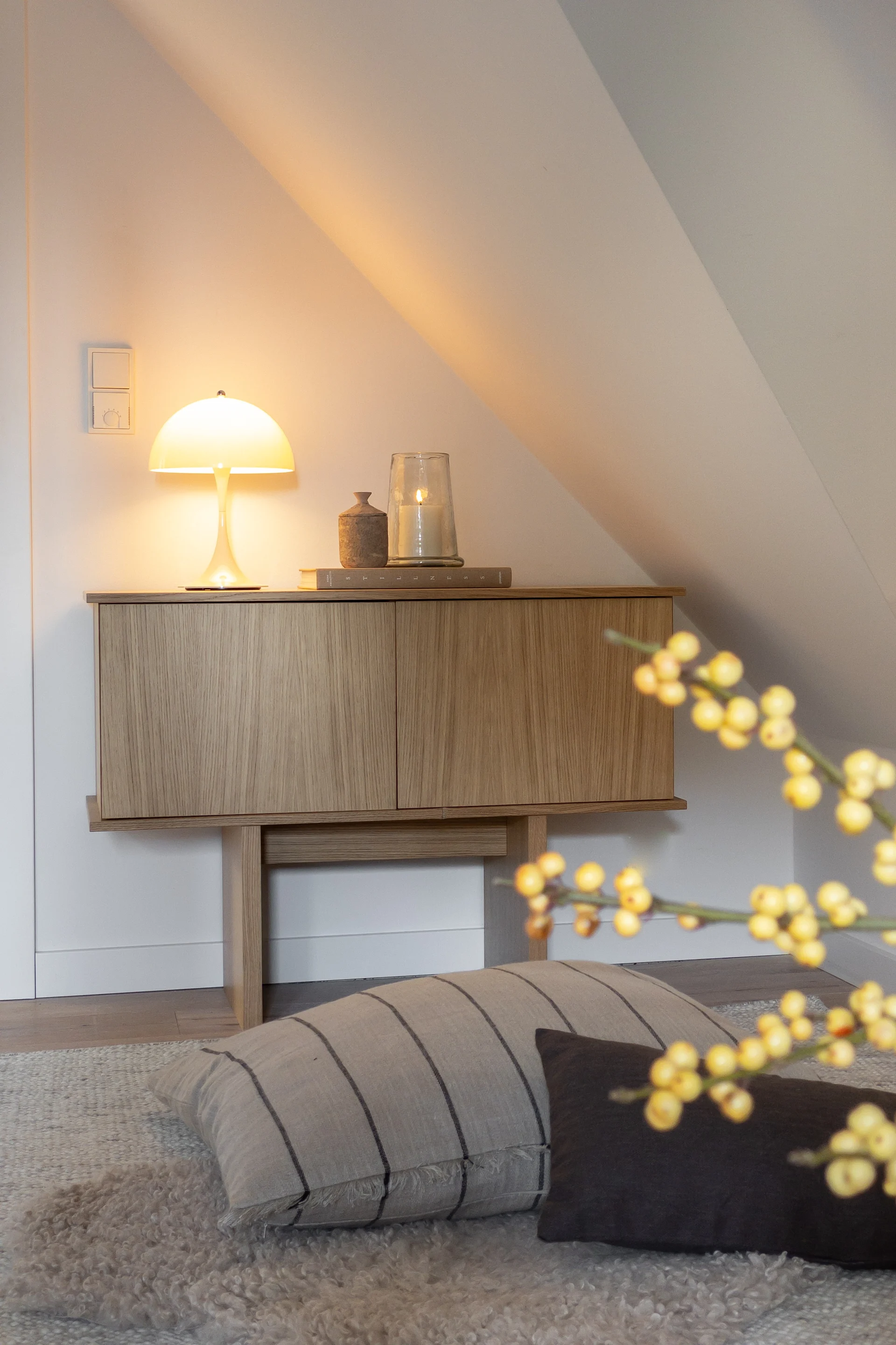 Un mueble de madera con una lámpara de hongo encendida y una vela, en una pared blanca e inclinada de una habitación acogedora.