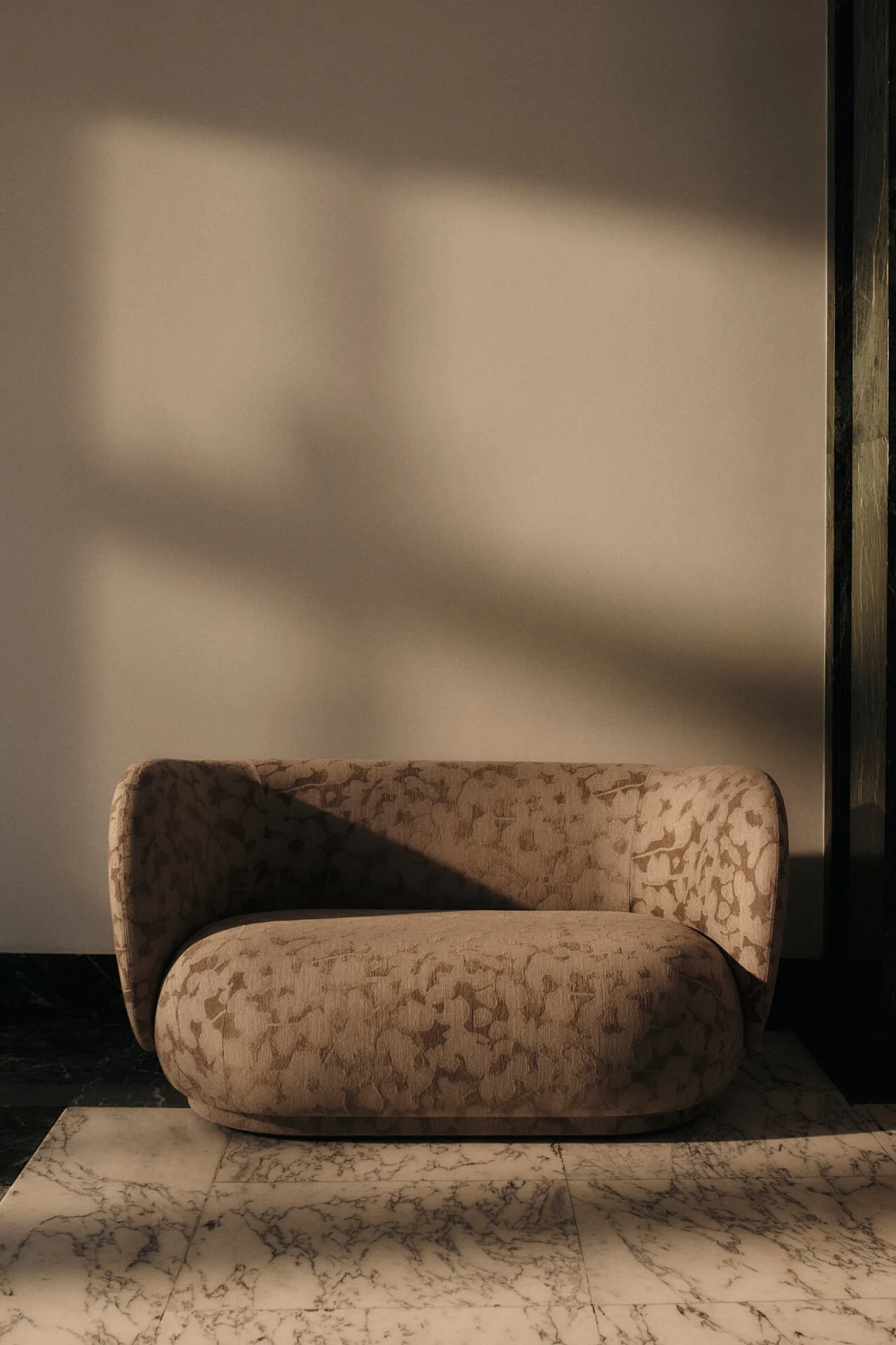 Sillón beige redondeado con tela de patrón sutil sobre suelo de mármol blanco, con sombras dramáticas por luz cálida.