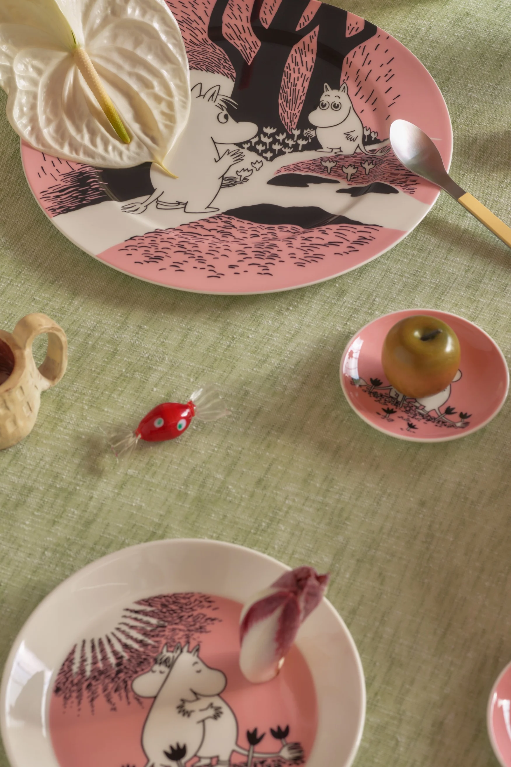 Mesa puesta con platos rosas de Moomin, flor de anturio, cuchara, manzana verde, caramelo rojo y una taza de madera sobre un mantel verde.