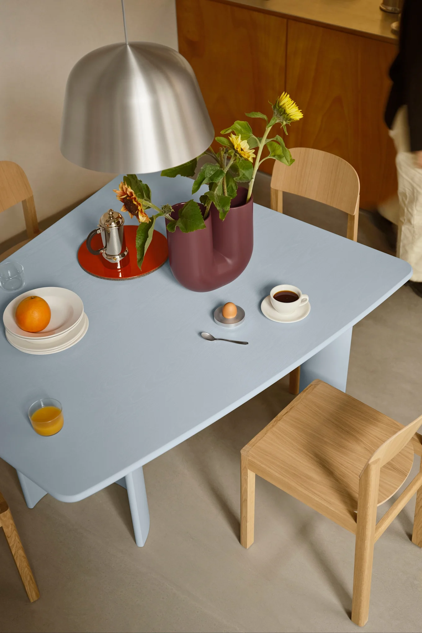 Una mesa de comedor azul puesta para el desayuno con café, zumo de naranja, un huevo y girasoles en un jarrón burdeos, bajo una lámpara plateada.