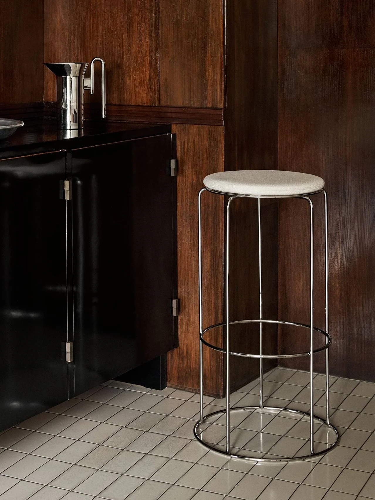 Un gabinete negro con utensilios de bar y un taburete cromado con asiento claro, sobre paneles de madera oscura y suelo de baldosas.