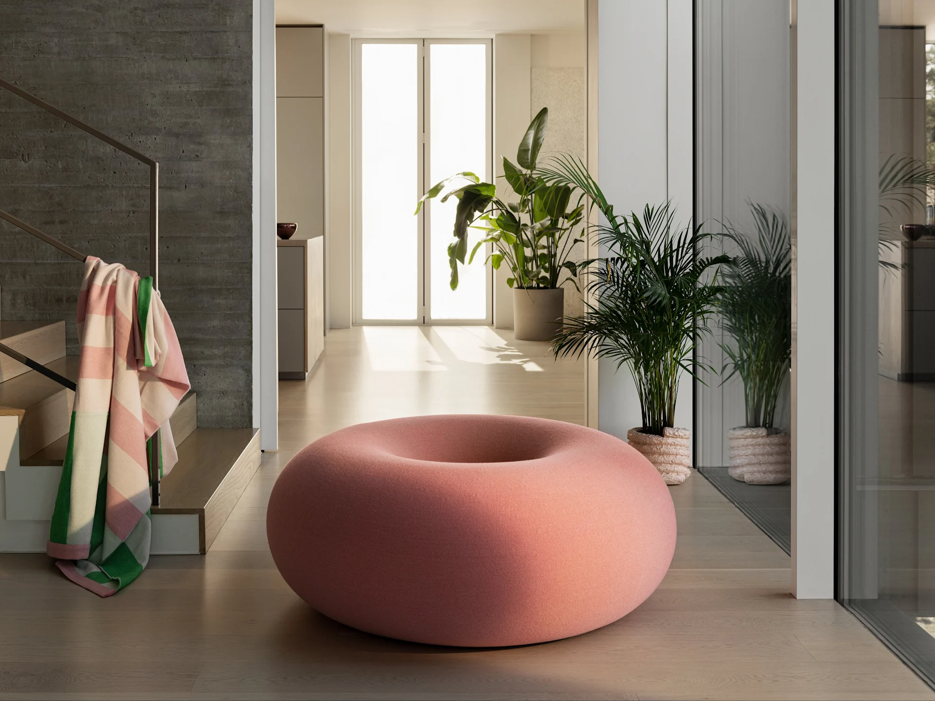 Un puf rosa grande con forma de donut sobre un suelo de madera clara en una habitación moderna con plantas y una pared de hormigón.