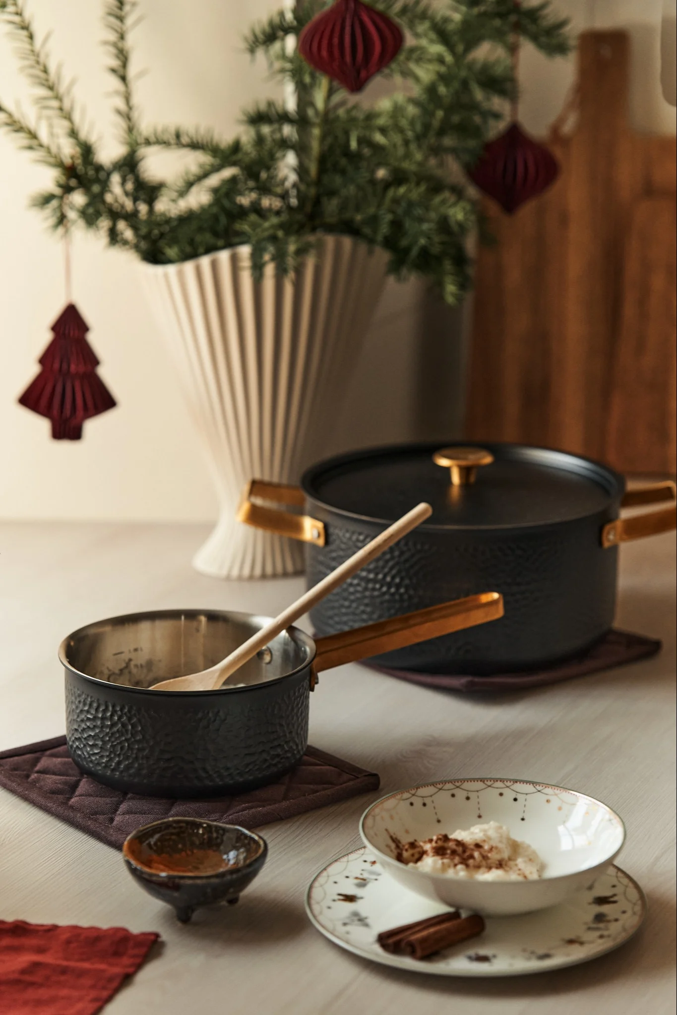 Un desayuno de Adviento preparado para el primer domingo de Adviento, con la cazuela “Viggo” de Vargen & Thor. Sobre la mesa, la cazuela está junto a un plato de arroz con leche, pan de jengibre y un cuenco; al fondo se ve un árbol de Navidad en un jarrón.