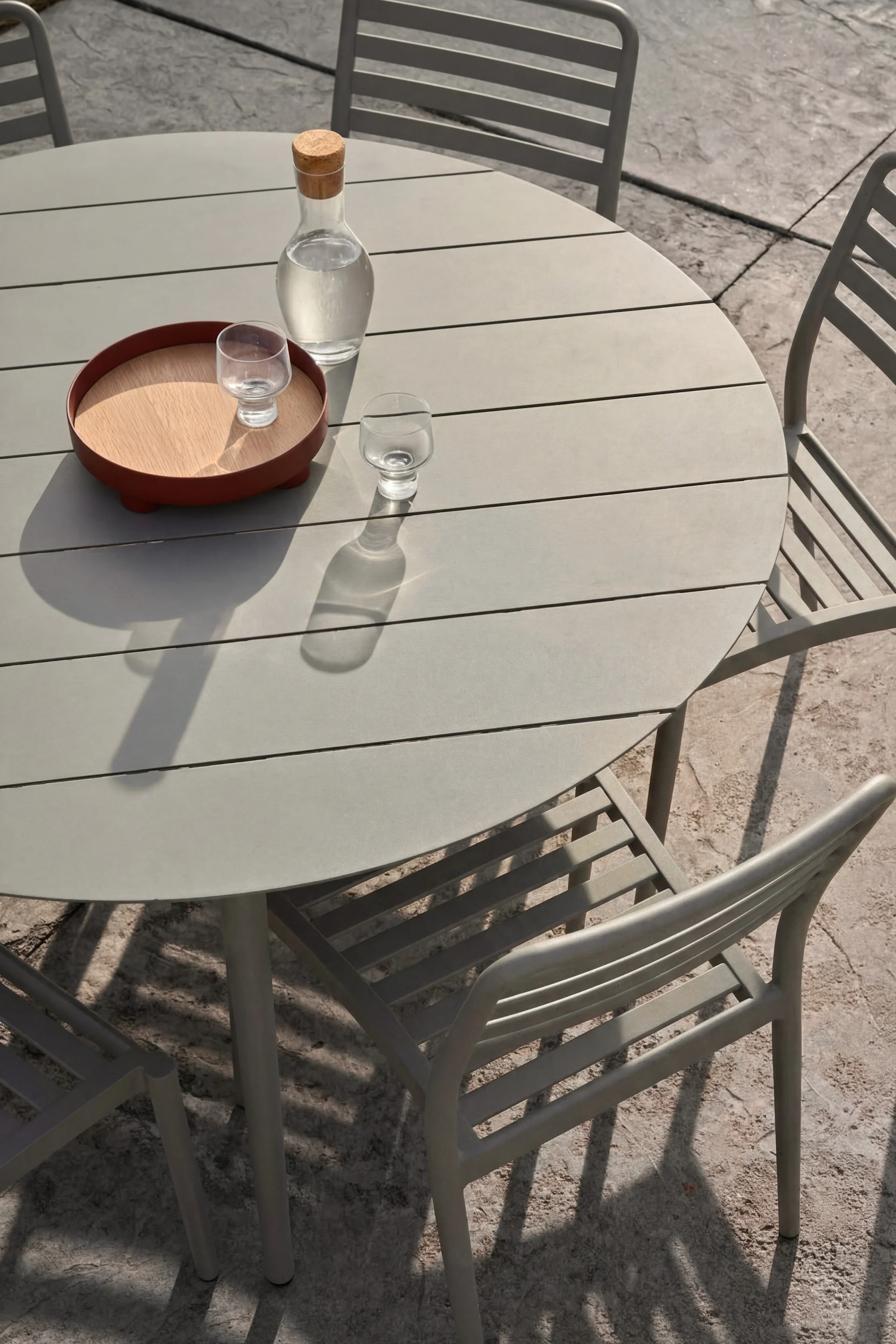 Mesa redonda gris de exterior, tres sillas. Jarra de agua, dos vasos (uno en bandeja de madera) en la mesa.
