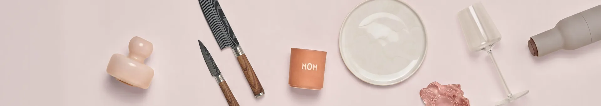 Cerámica rosa, dos cuchillos, taza 'MOM', plato blanco, cristal rosa, copa y botella gris sobre superficie rosa.