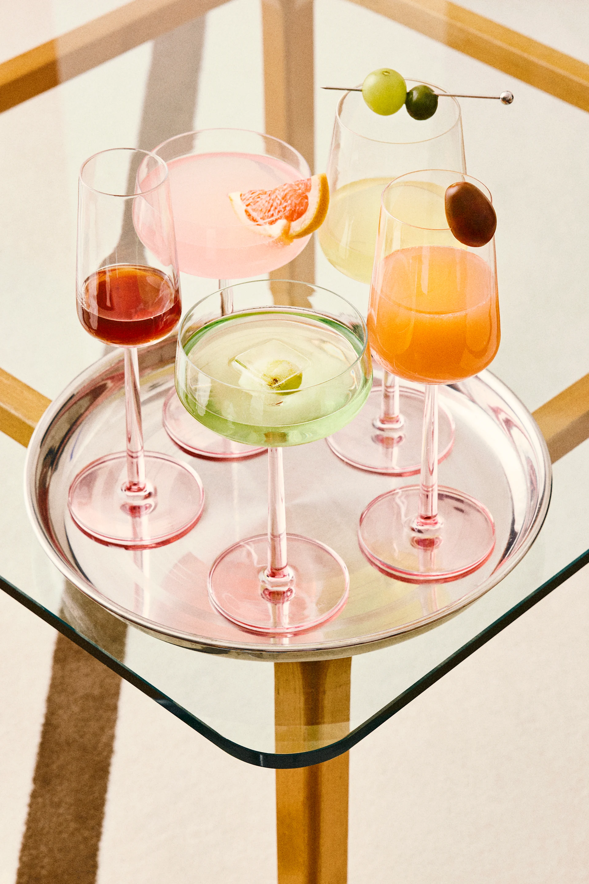 Cinco cócteles coloridos en una bandeja de plata, incluyendo bebidas rosas, verdes, naranjas y rojas con varias guarniciones, sobre una mesa de cristal.
