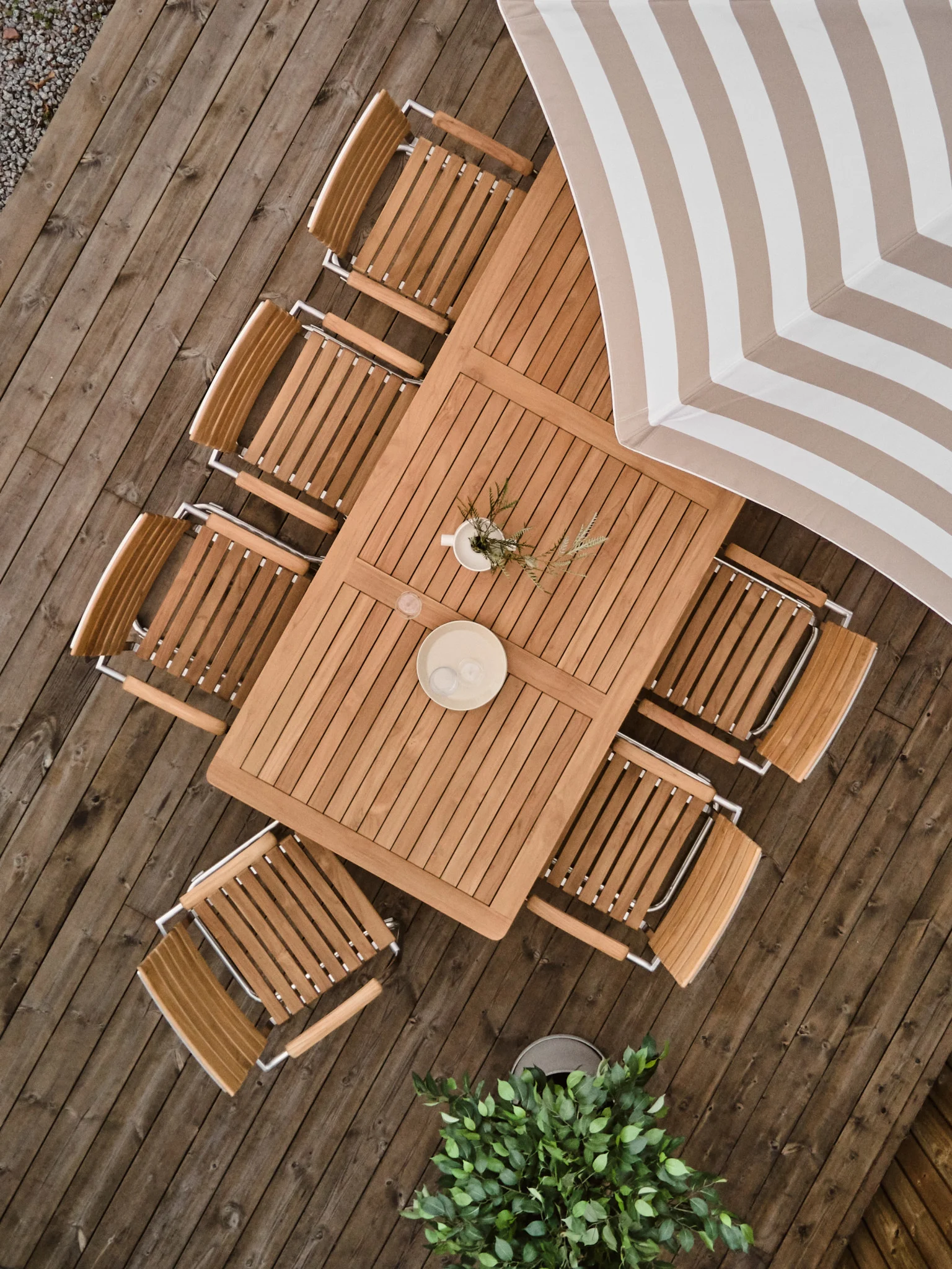Vista aérea de un conjunto de comedor de madera para exteriores en una terraza, con una mesa rectangular, ocho sillas y una sombrilla a rayas.