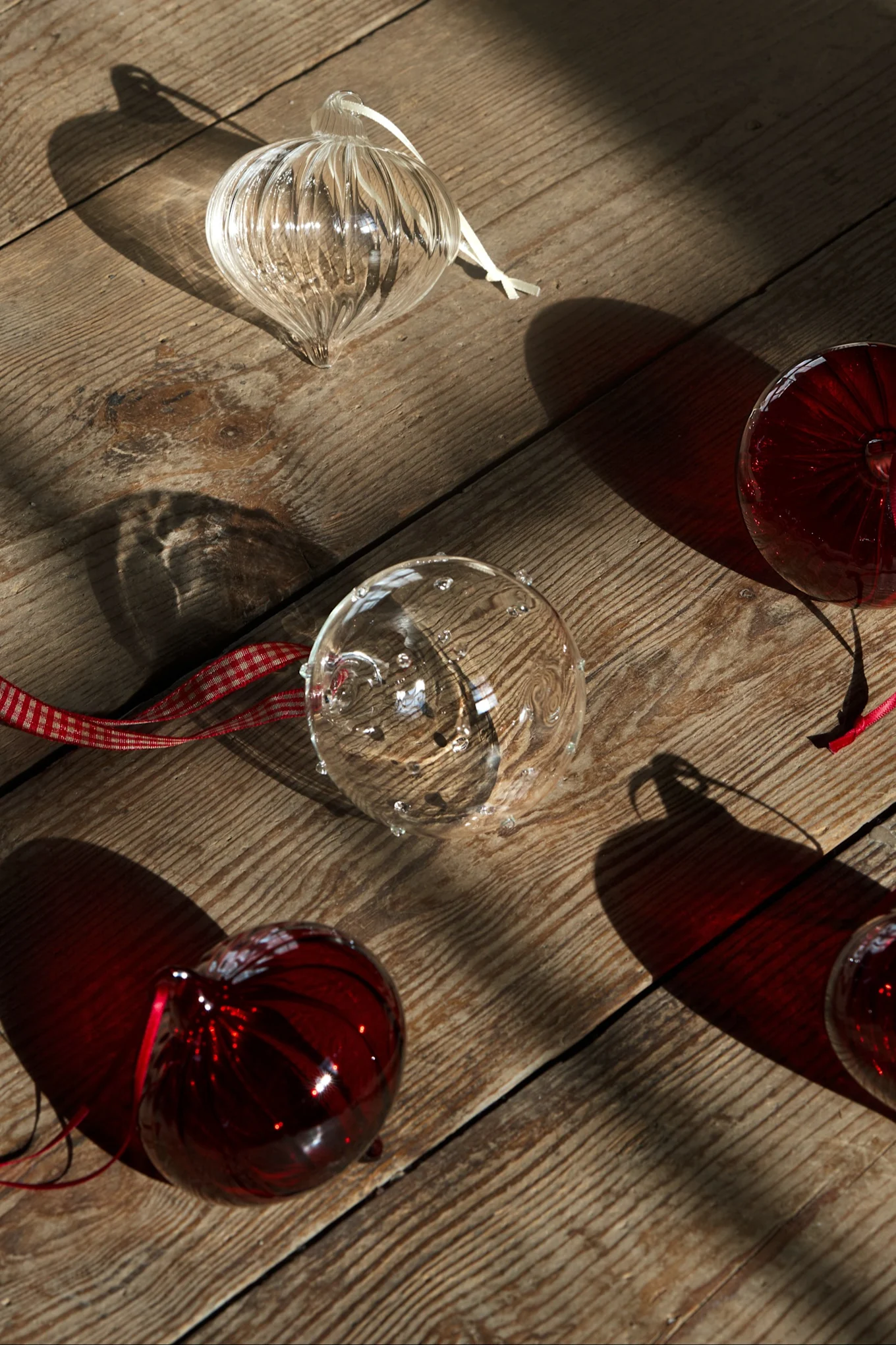 Adornos navideños de cristal transparentes y rojos con cintas esparcidos sobre una superficie de madera soleada, creando sombras.