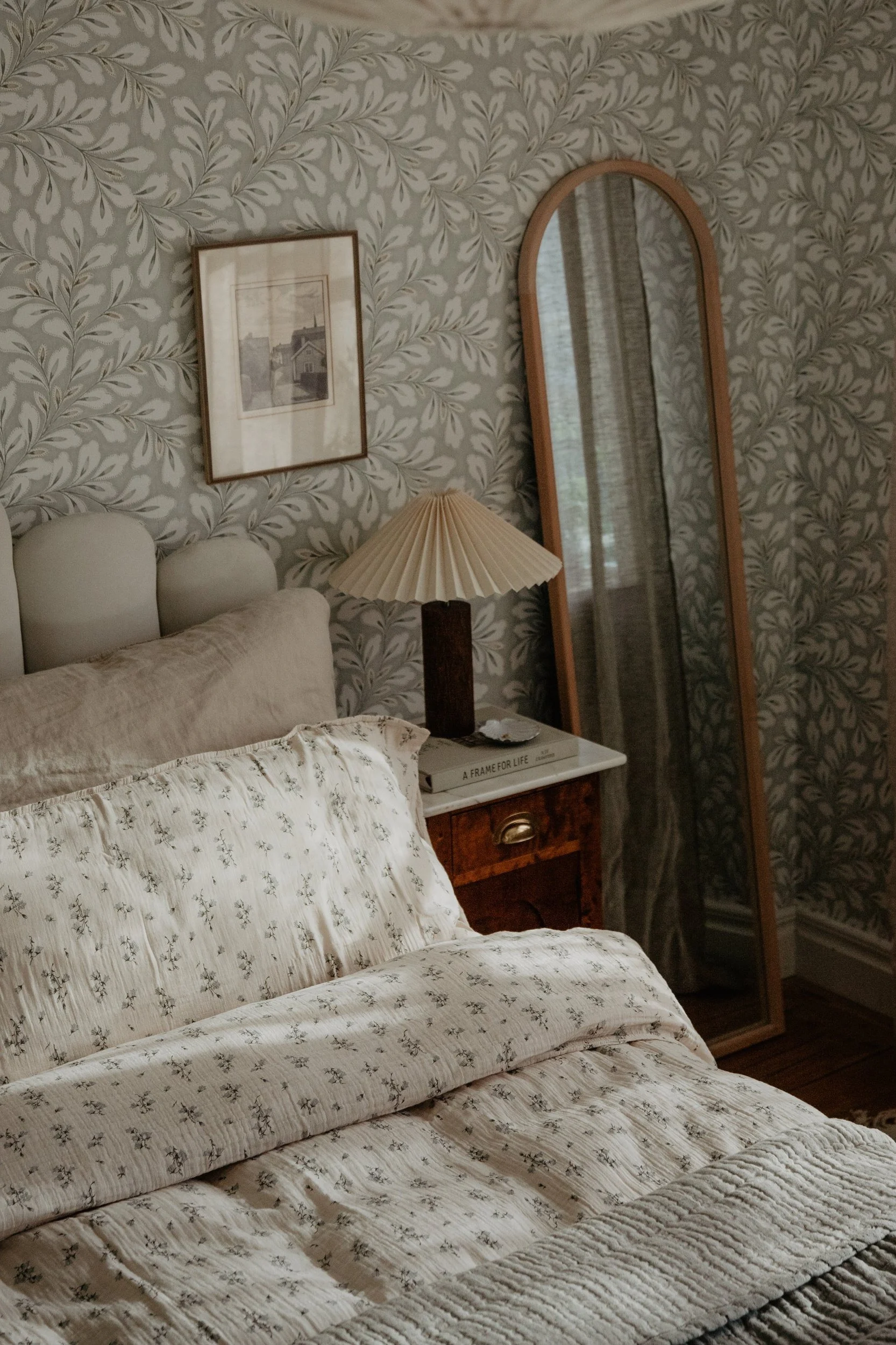 Un dormitorio con ropa de cama crema estampada, una lámpara plisada en una mesita de noche de madera y un espejo alto, sobre papel pintado de hojas.