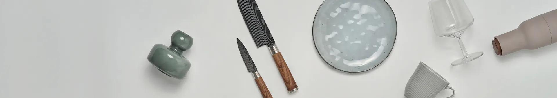 Varios artículos de cocina y comedor, incluyendo un jarrón verde, dos cuchillos, un plato moteado, una copa de vino, una botella beige y una taza estampada.